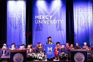 Student speaker at Commencement stage surrounded by faculty